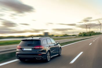 A dark-colored car drives along a highway surrounded by green fields and a warm sunset sky.